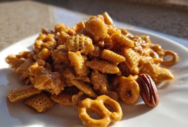 A mound of glossy, caramel-coated toffee chex mix recipe featuring cereal pieces, pretzels, and pecans on a white plate.
