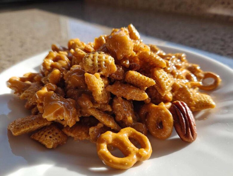 A mound of glossy, caramel-coated toffee chex mix recipe featuring cereal pieces, pretzels, and pecans on a white plate.