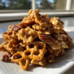 A close-up pile of sticky, glazed toffee chex mix recipe featuring pretzels and pecans, glistening in the sunlight.