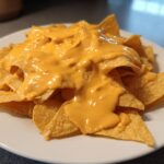 A white plate piled high with tortilla chips generously smothered in bright orange nacho cheese sauce.