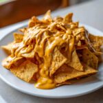 Close-up of tortilla chips piled high on a white plate, generously covered in thick, creamy nacho cheese sauce.