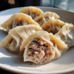 Close-up of steamed Traditional Chinese Dumplings on a white plate, one is cut open revealing the savory meat filling.