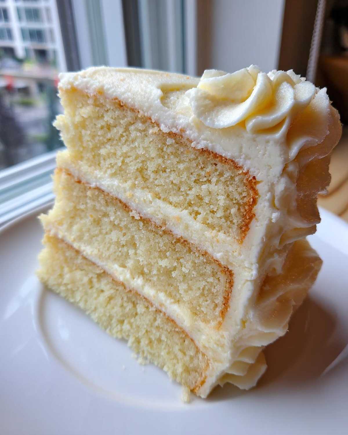 Close-up of a thick slice of moist vanilla cake recipe with three layers and creamy white frosting.