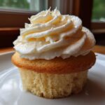 A single, moist vanilla cupcakes topped with a generous swirl of creamy white frosting, sitting on a white plate.
