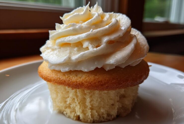 A single, moist vanilla cupcakes topped with a generous swirl of creamy white frosting, sitting on a white plate.