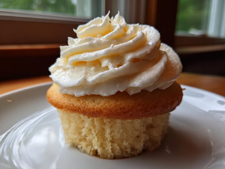 A single, moist vanilla cupcakes topped with a generous swirl of creamy white frosting, sitting on a white plate.