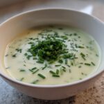A close-up of a white bowl filled with creamy, light green leek soup, topped generously with fresh chopped chives.
