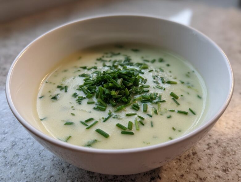 A close-up of a white bowl filled with creamy, light green leek soup, topped generously with fresh chopped chives.