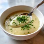 A spoonful of creamy, velvety leek soup topped with fresh green chives being lifted from a white bowl.