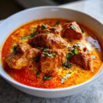 Close-up of vibrant, orange-red butter chicken pieces swimming in a rich, creamy sauce, garnished with fresh cilantro.