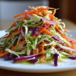 A tall mound of colorful, crunchy cabbage salad featuring shredded green cabbage, carrots, and red onion on a white plate.