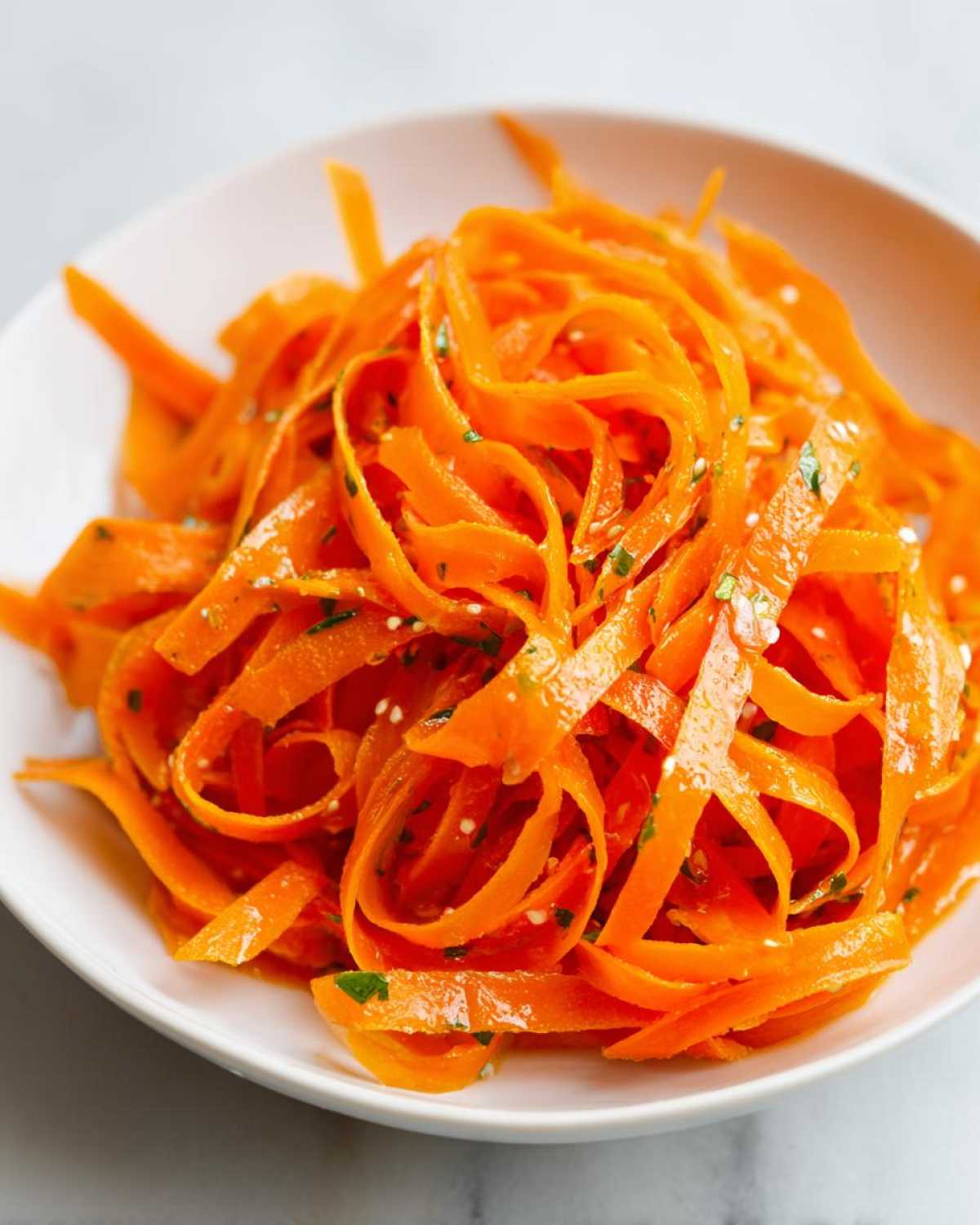 Close-up of bright orange raw carrot salad ribbons tossed with herbs and sesame seeds in a white bowl.