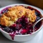 Close-up of a warm mixed berry cobbler with a golden, crumbly topping and bubbling purple fruit filling, served in a white ramekin with a spoon.