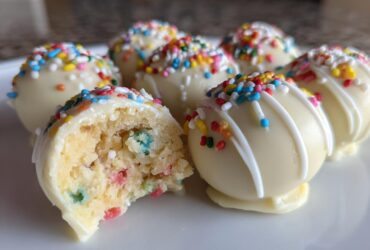 A close-up of several white chocolate-coated sugar cookie truffles, one cut open revealing colorful sprinkles inside.