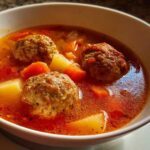 Close-up of a white bowl filled with rich, red broth albondigas soup featuring three large meatballs, potatoes, and carrots.