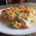 Close-up of a hearty serving of chicken noodle casserole, showing shredded chicken, egg noodles, peas, carrots, and a bubbly, golden cheese topping.