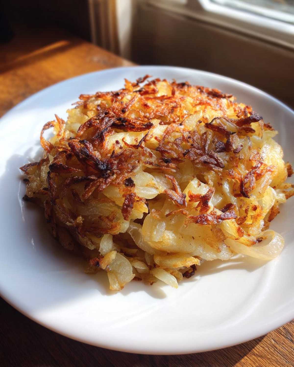 A golden-brown, crispy mound of fried potatoes served on a white plate, catching bright sunlight.