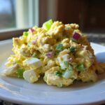 A mound of creamy honey mustard chicken salad mixed with chopped celery, red onion, and hard-boiled egg pieces on a white plate.