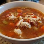 A close-up view of a hearty bowl of amazing maryland crab soup, featuring large chunks of white crab meat and diced vegetables in a rich, reddish broth.