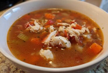 A close-up view of a hearty bowl of amazing maryland crab soup, featuring large chunks of white crab meat and diced vegetables in a rich, reddish broth.