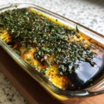 Close-up of a glass dish containing an amazing olive oil bread dip topped with fresh green herbs, spices, and dark balsamic vinegar.