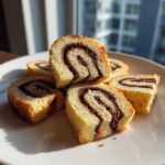Several slices of light sponge cake pinwheel cookies with a dark chocolate swirl stacked on a white plate.