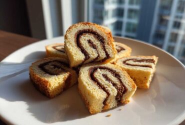 Several slices of light sponge cake pinwheel cookies with a dark chocolate swirl stacked on a white plate.
