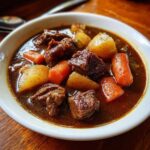 Close-up of a bowl filled with rich, dark brown pork stew featuring tender chunks of meat, potatoes, and bright orange carrots.