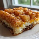 A thick slice of tater tot casserole showing layers of ground meat, creamy filling, cheese, and a crispy tater tot topping.