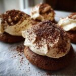 Close-up of rich, dark chocolate tiramisu cookies topped with a swirl of cream and dusted heavily with cocoa powder.