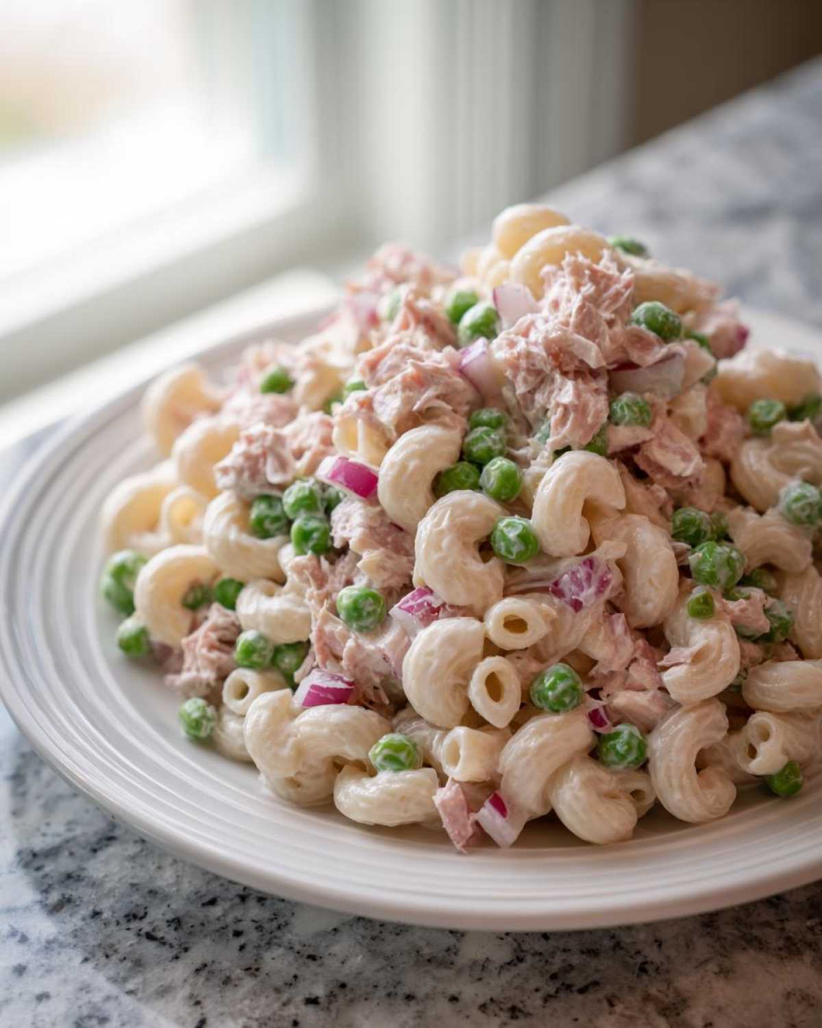A generous serving of creamy tuna pasta salad mixed with elbow macaroni, bright green peas, and diced red onion on a white plate.