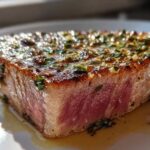 Close-up of a perfectly seared tuna steak, showing a pink, rare center and a glistening herb and garlic crust.