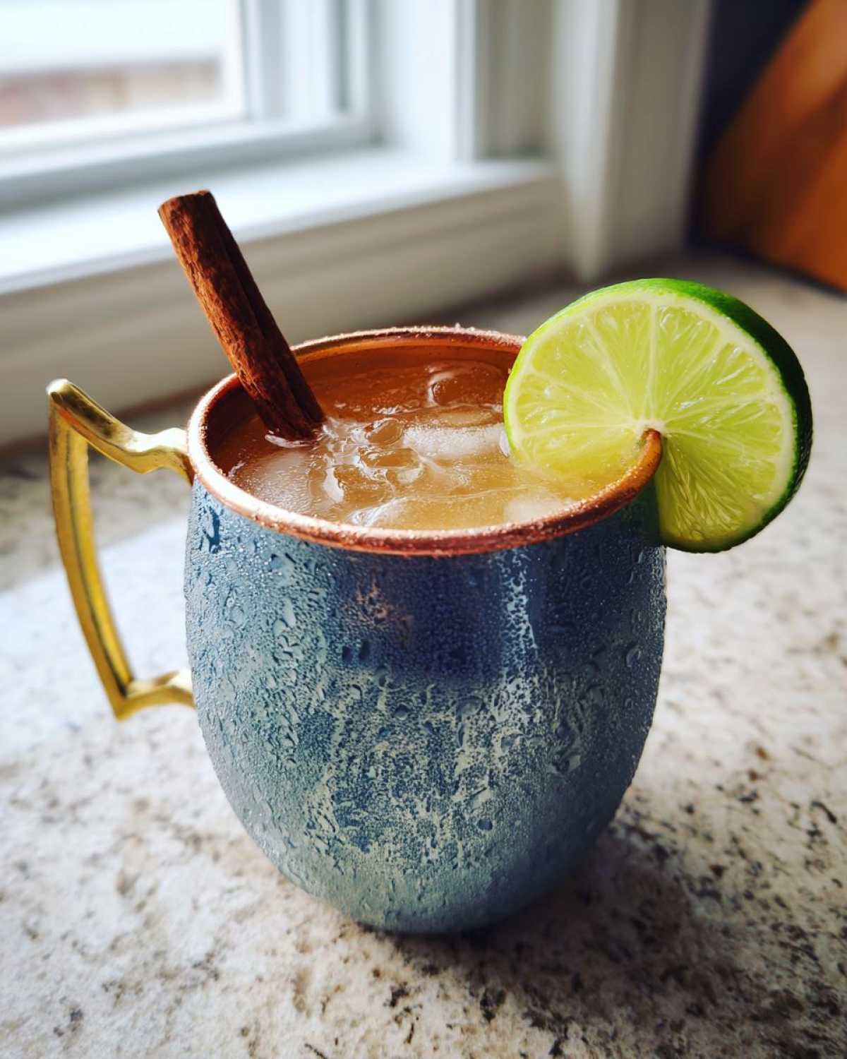 A condensation-covered blue copper mug filled with an apple cider Moscow mule, garnished with a lime wheel and cinnamon stick.