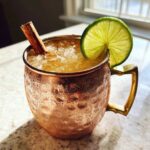 A frosty copper mug filled with an apple cider Moscow mule, garnished with a lime wheel and cinnamon stick.