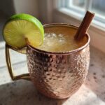 A refreshing apple cider moscow mule served over crushed ice in a hammered copper mug, garnished with a lime wheel and cinnamon stick.