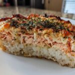 A thick slice of baked salmon sushi bake showing layers of white rice, pink salmon filling, and a browned, seasoned topping.
