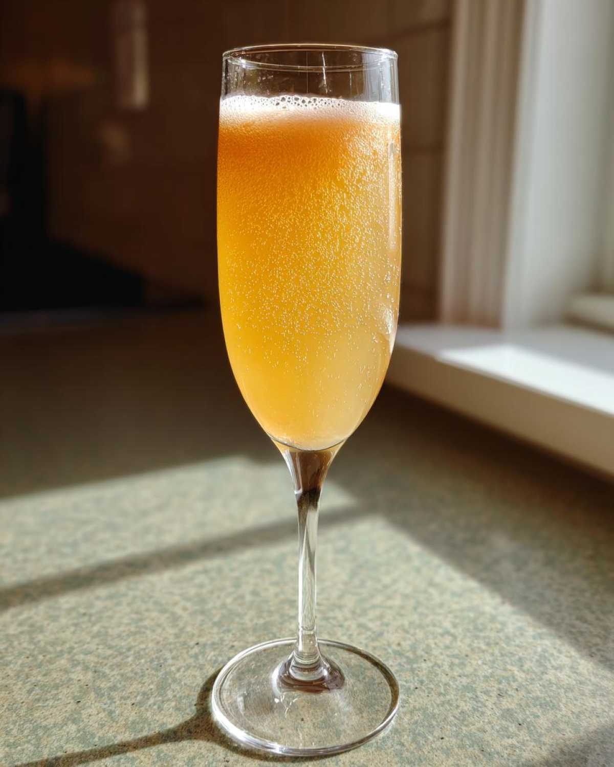 A tall, fizzy bellini recipe cocktail served in a champagne flute glass, catching sunlight on a countertop.
