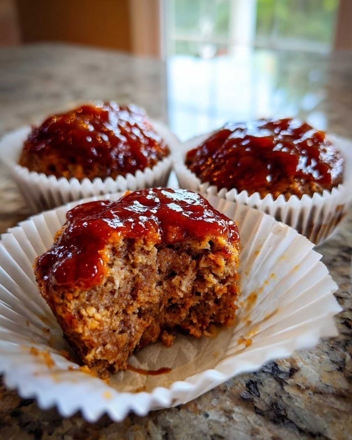 Close-up of a delicious mini meatloaf with a bite taken out, showing the texture and thick glaze, served in a white paper liner.