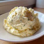 A close-up of a generous scoop of creamy boursin mashed potatoes mixed with visible green herbs, served on a white plate.