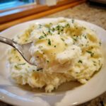 A spoonful of creamy boursin mashed potatoes topped with melted butter and fresh parsley, lifted from a white bowl.