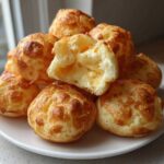 A stack of golden-brown Brazilian cheese bread, with one piece broken open revealing a gooey, cheesy interior.