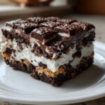 A rich, layered slice of brownie refrigerator cake featuring chocolate, white cream, and a chocolate bar topping.