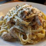 Close-up of a rich serving of chicken fettuccine alfredo, coated in creamy sauce and topped with grated cheese and herbs.