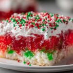 Close-up of a moist slice of christmas poke cake showing layers of white cake, red and green gelatin, whipped topping, and holiday sprinkles.