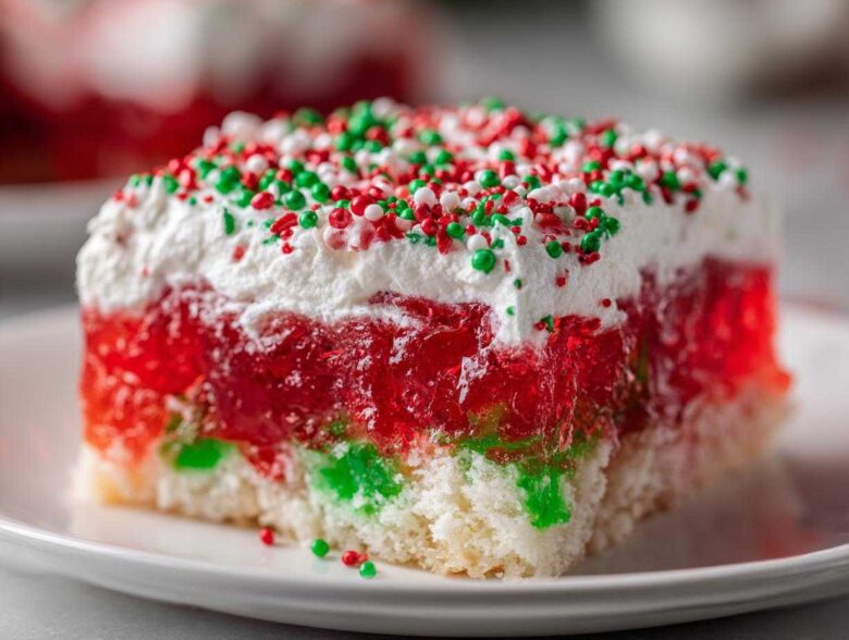 Close-up of a moist slice of christmas poke cake showing layers of white cake, red and green gelatin, whipped topping, and holiday sprinkles.