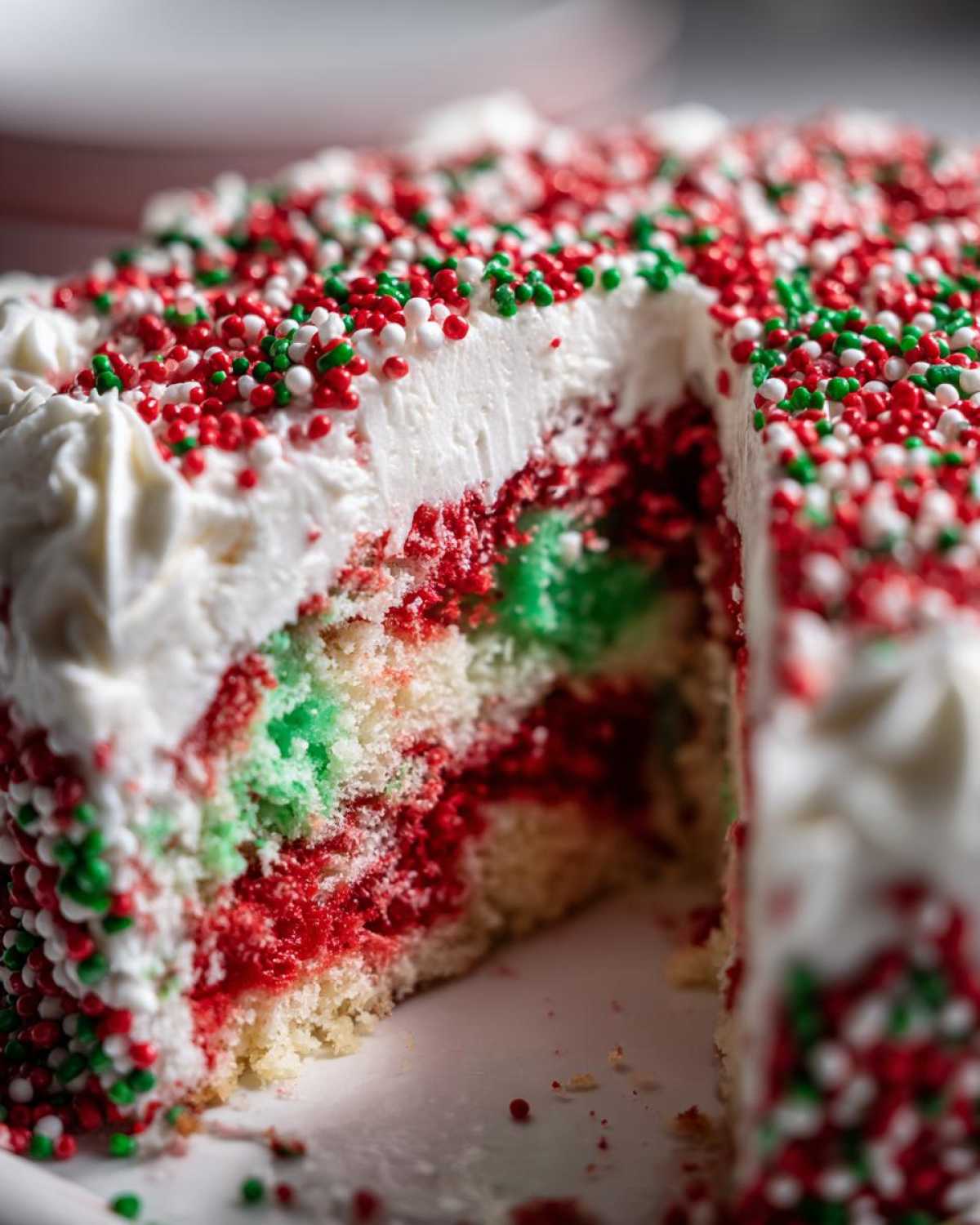 Close-up of a slice removed from a moist Christmas poke cake showing red and green layers and white frosting covered in holiday sprinkles.