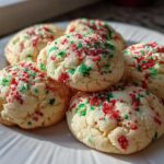 A close-up of five soft, round christmas sprinkle sugar cookies topped with red and green holiday sprinkles on a white plate.