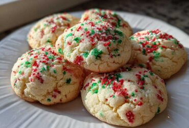 A close-up of five soft, round christmas sprinkle sugar cookies topped with red and green holiday sprinkles on a white plate.