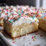 A thick slice of Christmas sugar cookie bars topped with white frosting and colorful sprinkles.