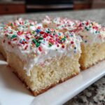 Close-up of four squares of fluffy Christmas sugar cookie bars topped with white frosting and colorful sprinkles.